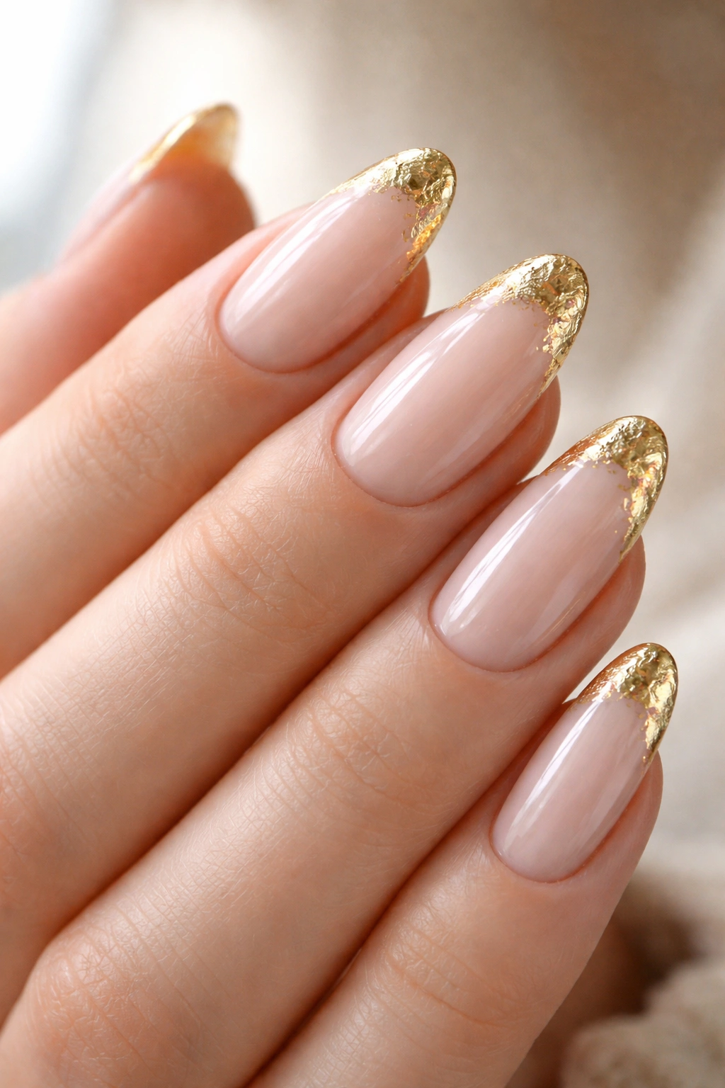 Close-up of almond nails with gold foil accents on the tips in a neutral studio setting