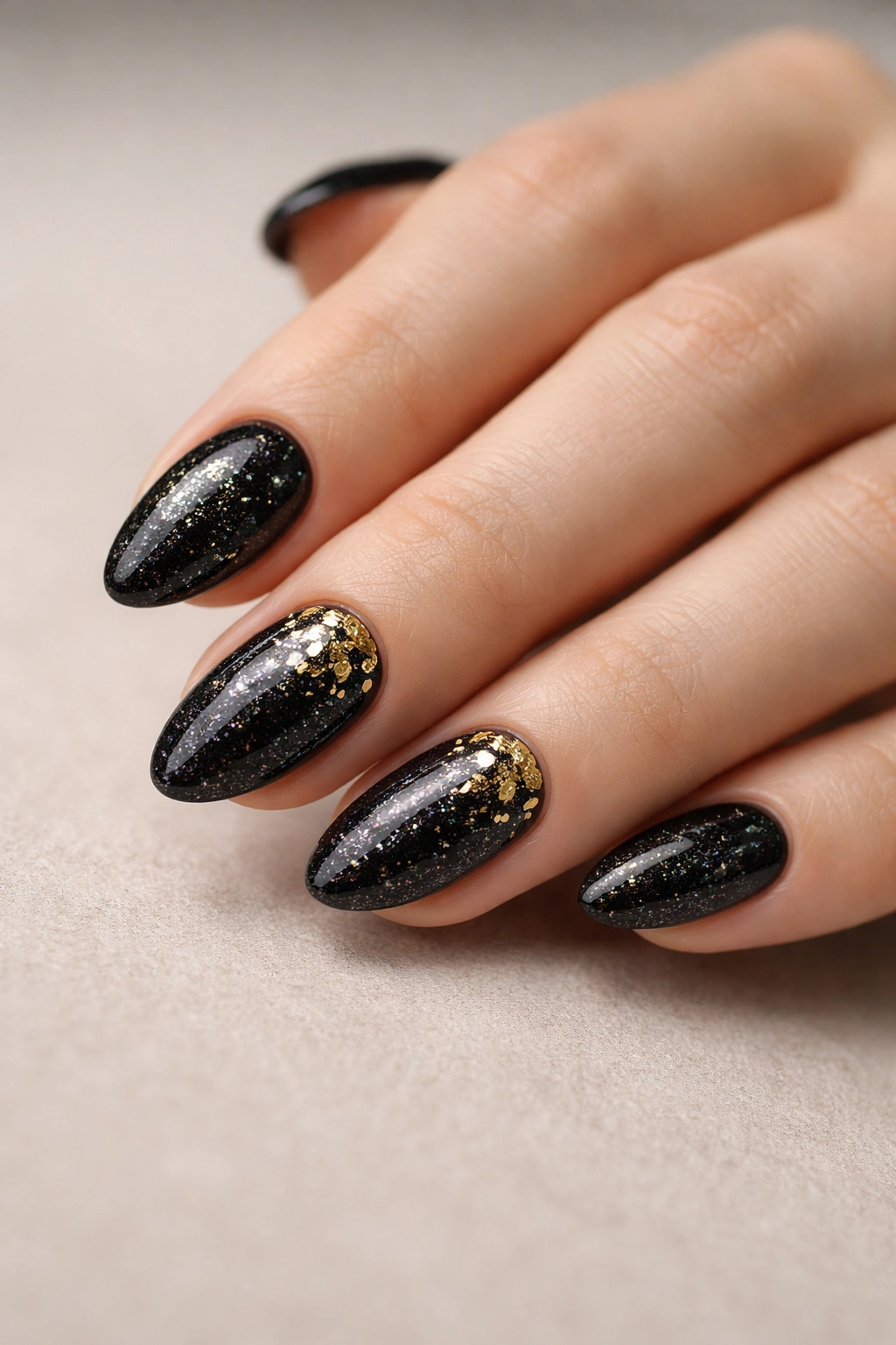 Close-up of short almond nails with a black glitter base and gold foil accents.
