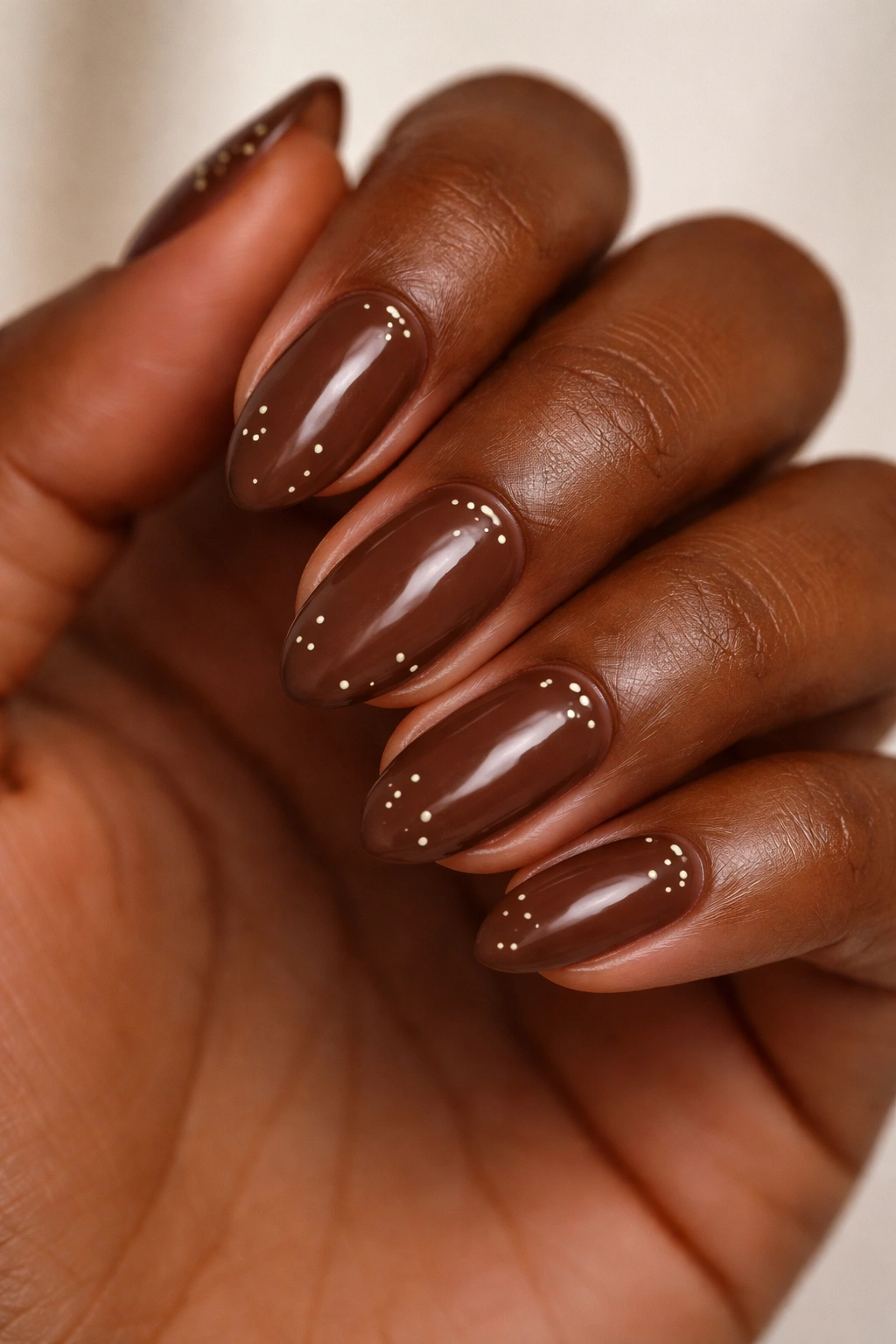 Close-up of a short almond nail with a rich brown polish and tiny cream and gold dots.