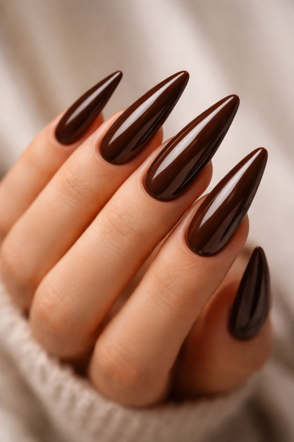 Close-up of glossy classic chocolate brown stiletto nails on a hand against a neutral background