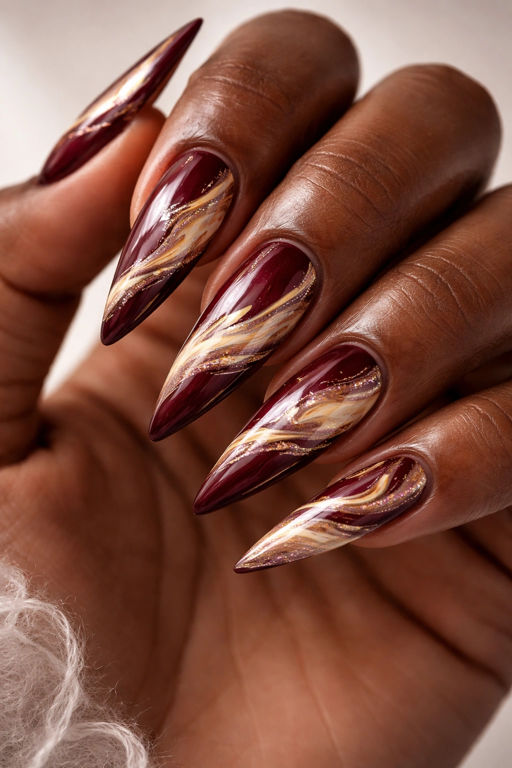 Burgundy stiletto nails with cream and gold marble swirls and glossy finish.