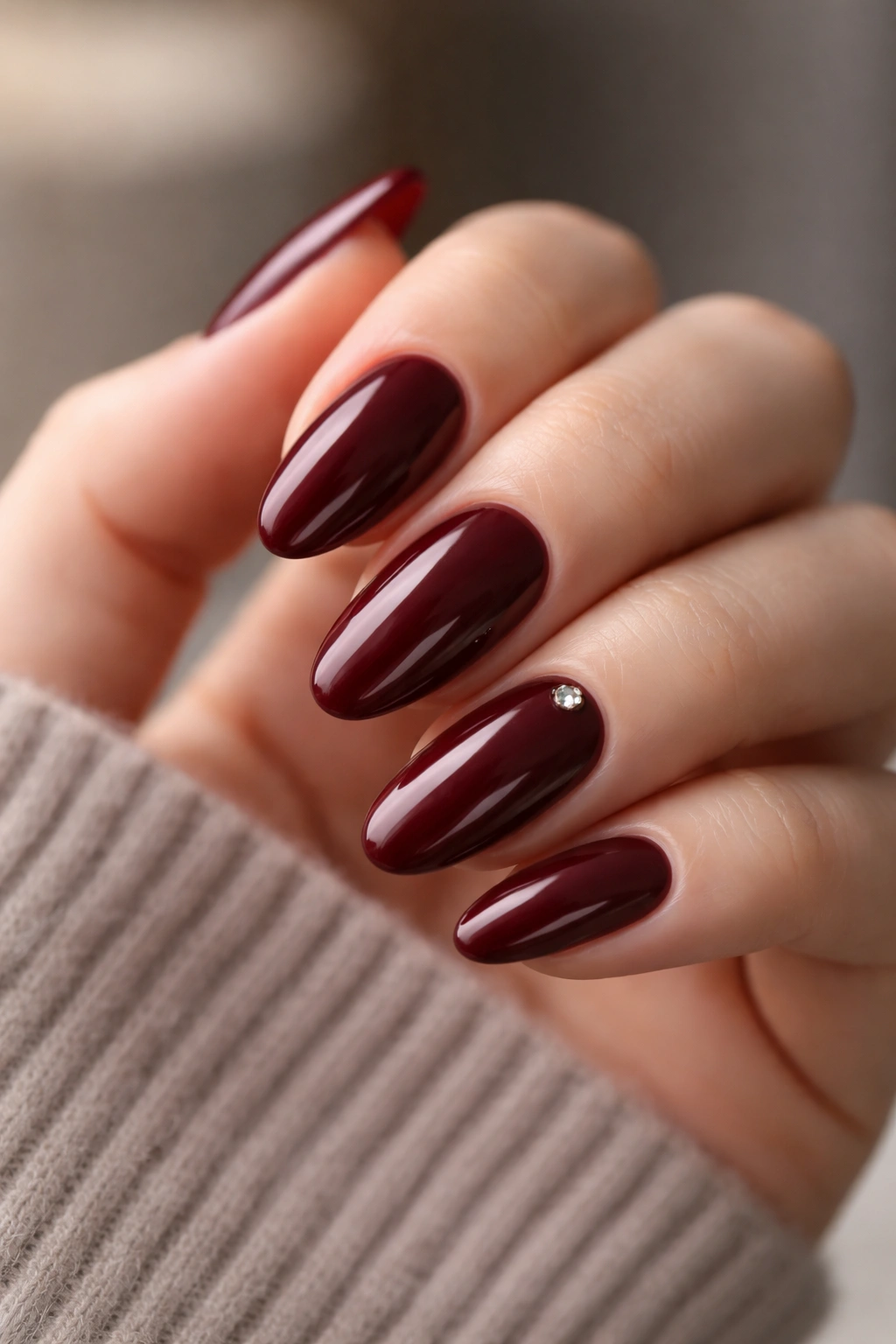 Close-up of burgundy long oval nails with a single crystal accent on one nail against a neutral background