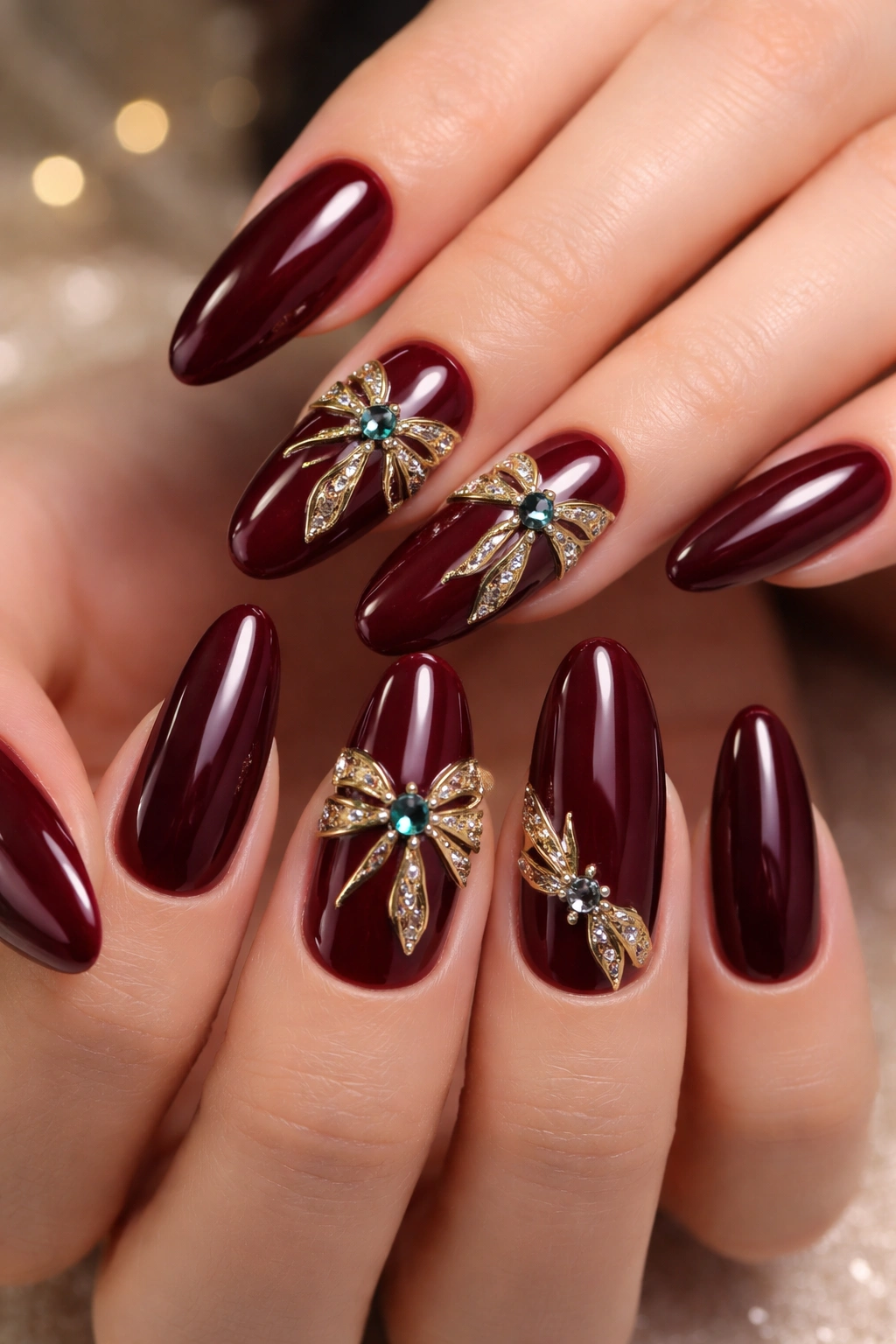 Burgundy nails with metallic gold bow and jewel accents on long oval nails in a studio close-up