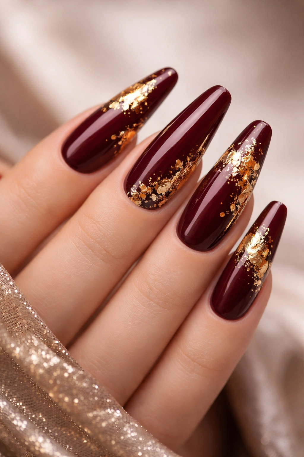 Close-up of burgundy long nails with irregular gold foil accents on a neutral background