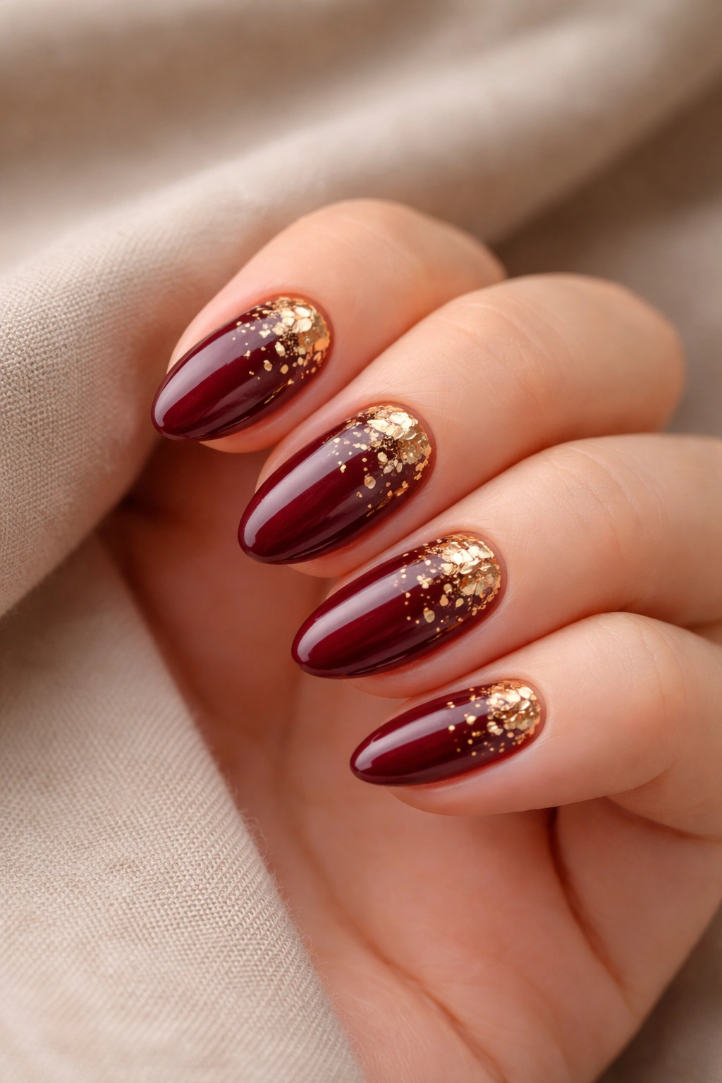 Close-up of short almond nails with deep burgundy and gold foil flakes