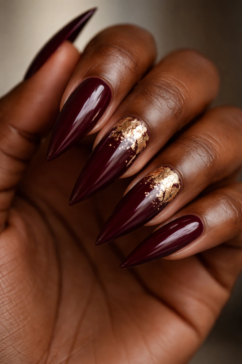 Close-up of deep burgundy stiletto nails with gold leaf accents on ring and middle fingers.