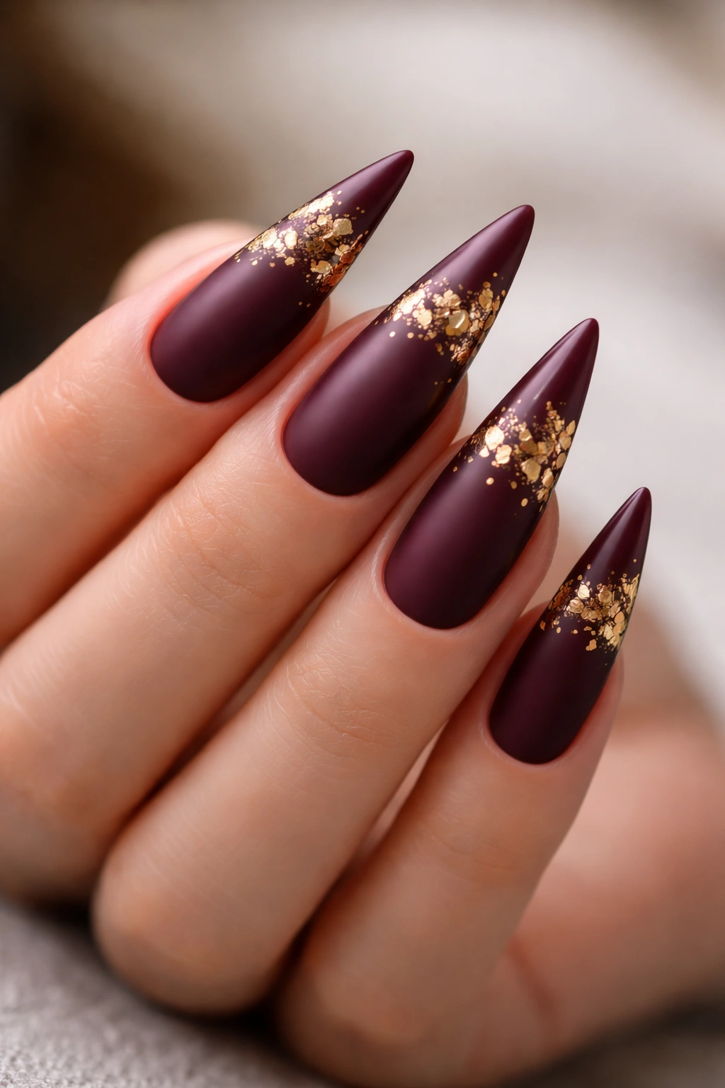 Close-up of burgundy stiletto nails with gold leaf accents near the tips.