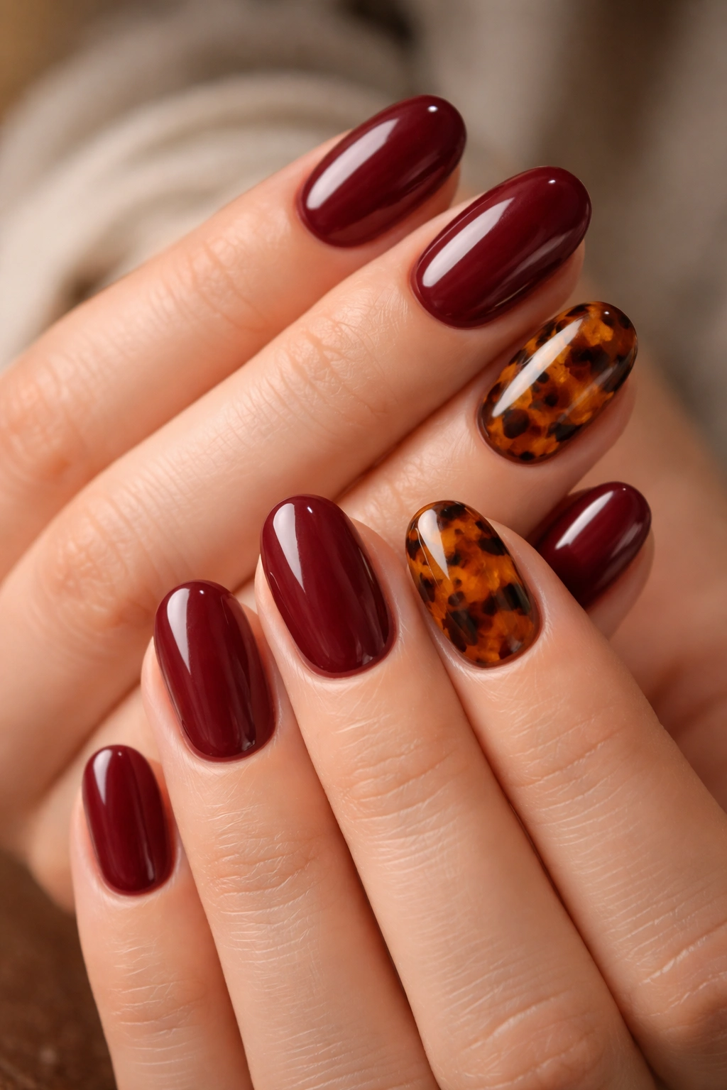 Close-up of burgundy short oval nails with tortoiseshell accents on a neutral background.