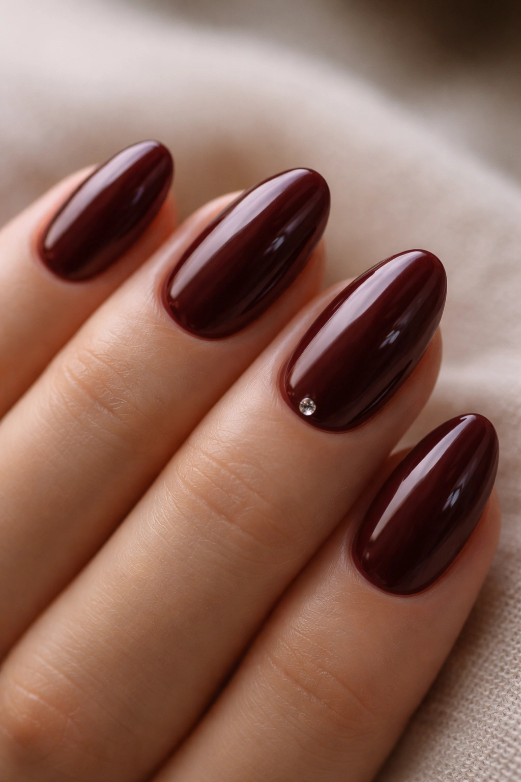 Short burgundy almond nails with a tiny rhinestone near the cuticle