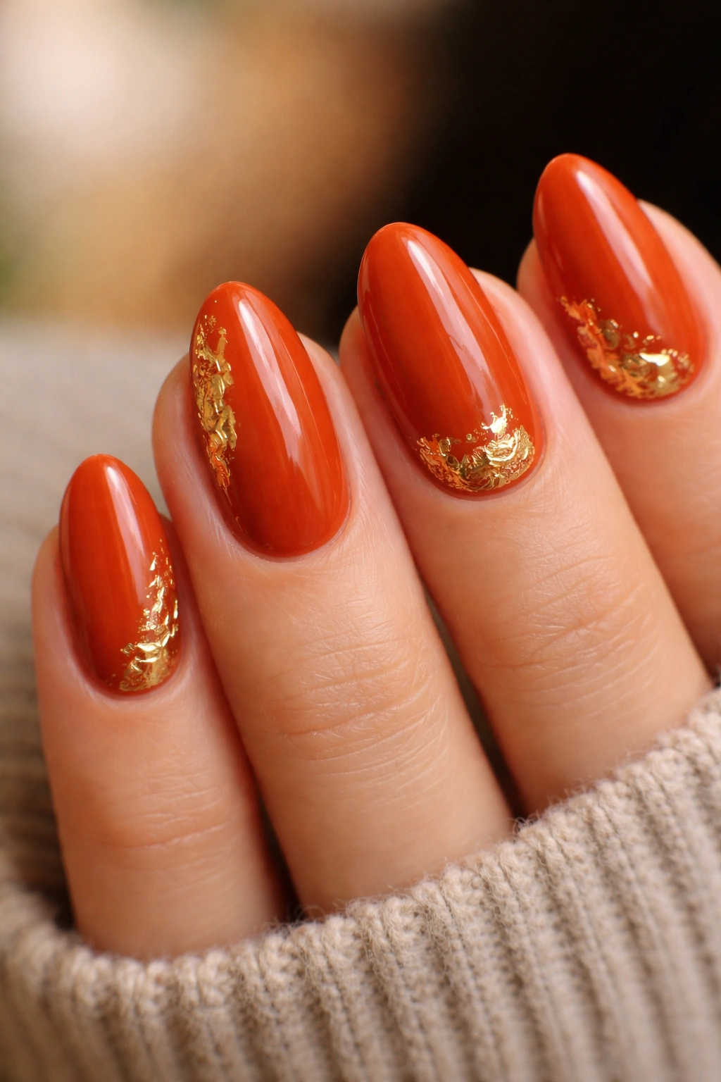 Short almond nails in burnt orange with gold foil accents along the cuticle and side.