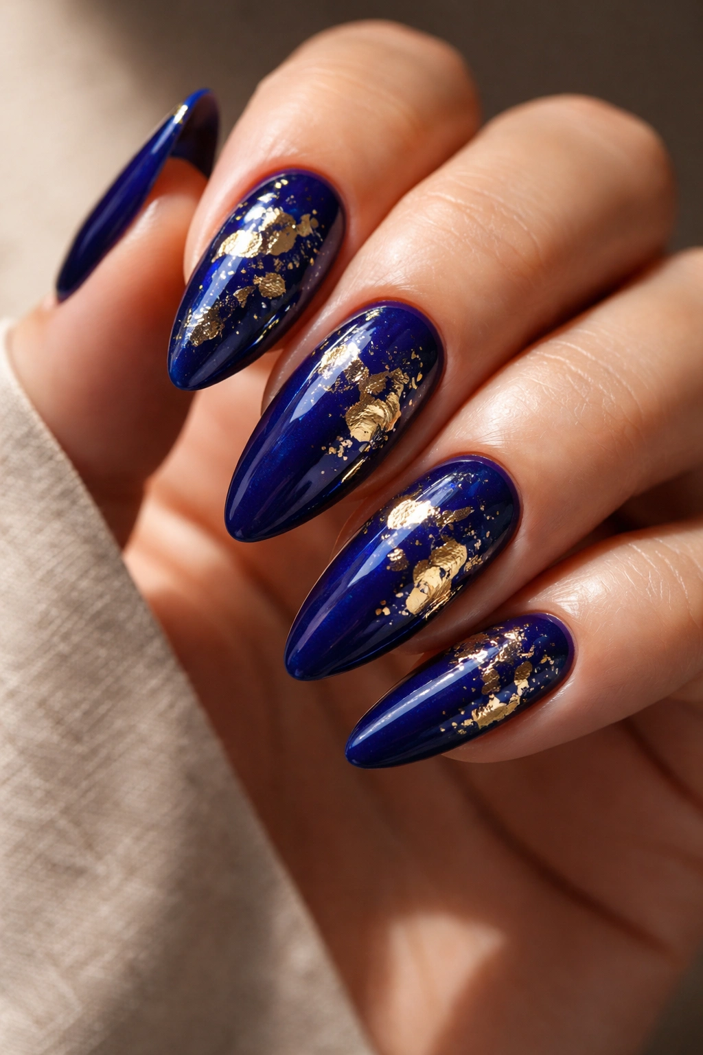 Close-up of almond nails in deep cobalt blue with gold foil accents on a neutral background