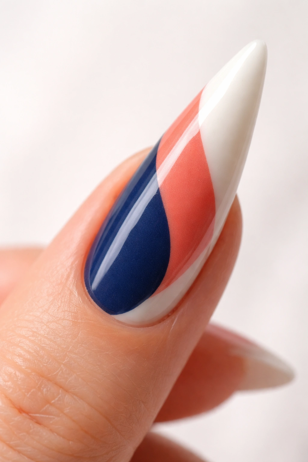 Close-up of a stiletto nail with curved color-block sections in navy, coral, and cream on a nude base