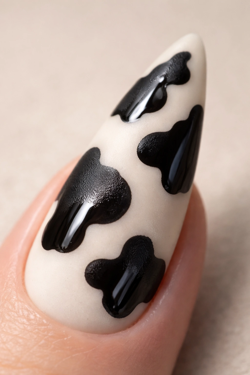 Close-up of a matte cow print stiletto nail with glossy patches on a cream base