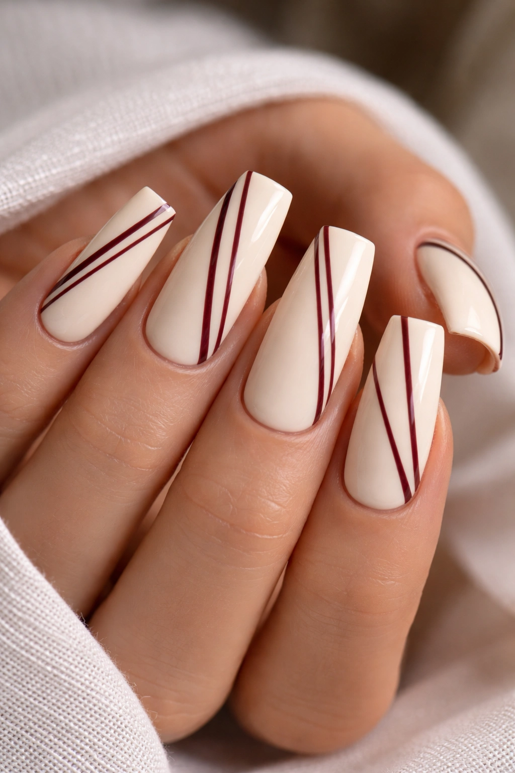Cream nails with thin burgundy stripes on a coffin shape