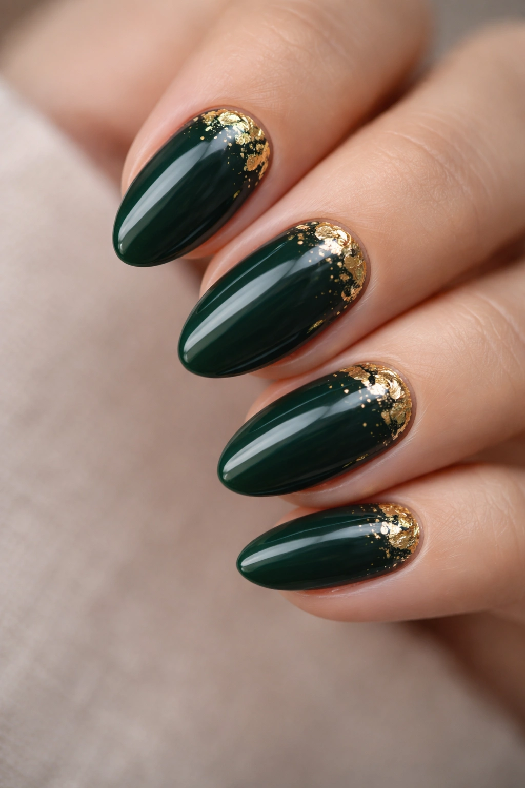 Close-up of short almond nails with dark green polish and gold foil accents.