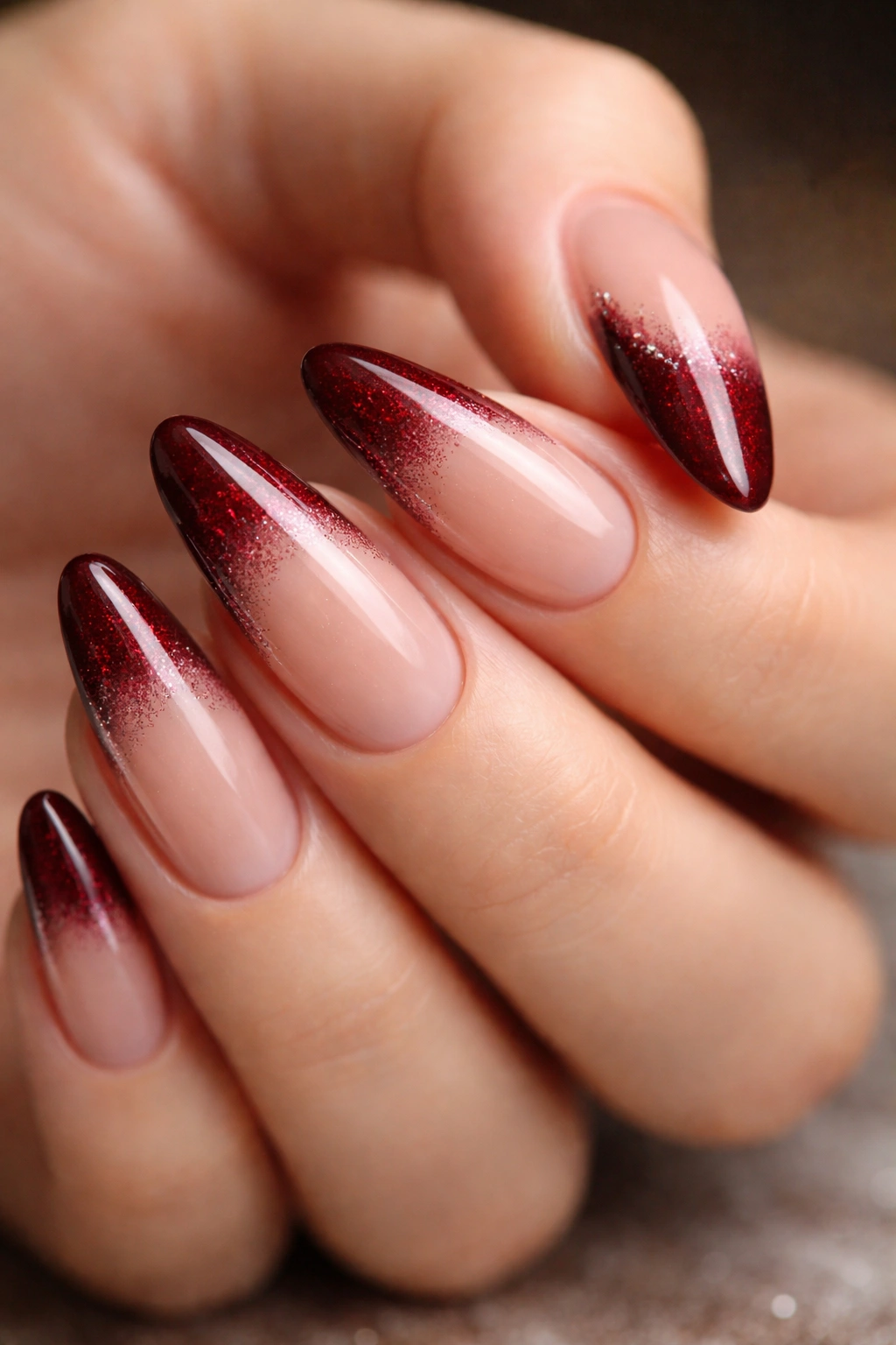 Close-up of almond nails with dark red tips and burgundy glitter gradient on nude base