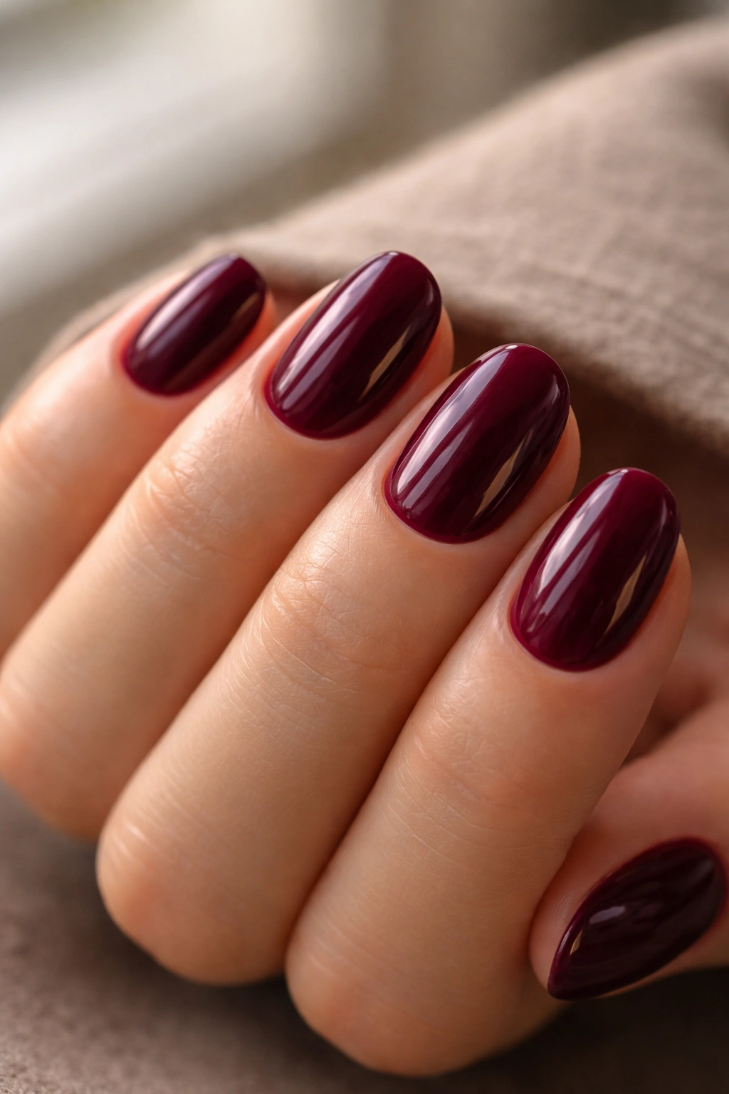 Close-up of deep berry short oval nails with glossy top coat on a neutral background