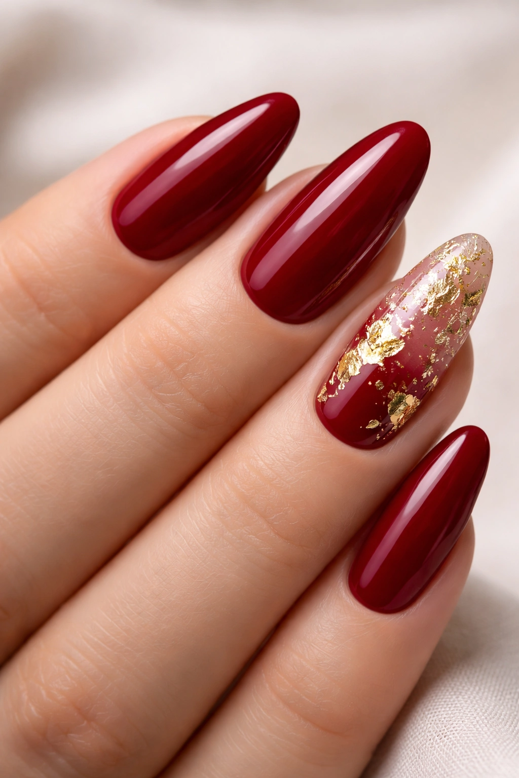 Close-up of long oval nails with deep red base and a gold foil accent nail
