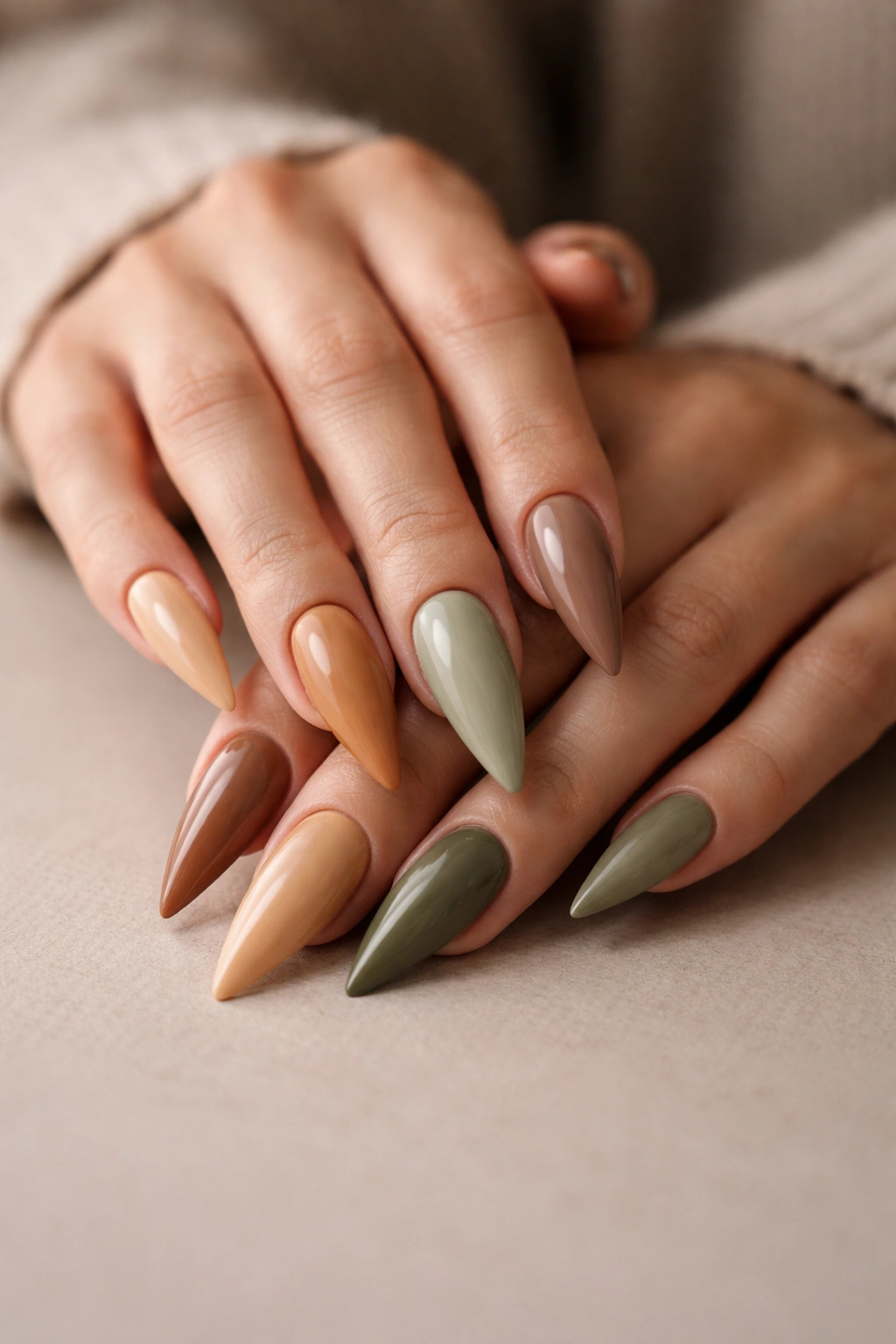 Close-up of earth-tone stiletto nails in caramel, sage, taupe on hands resting on a neutral surface