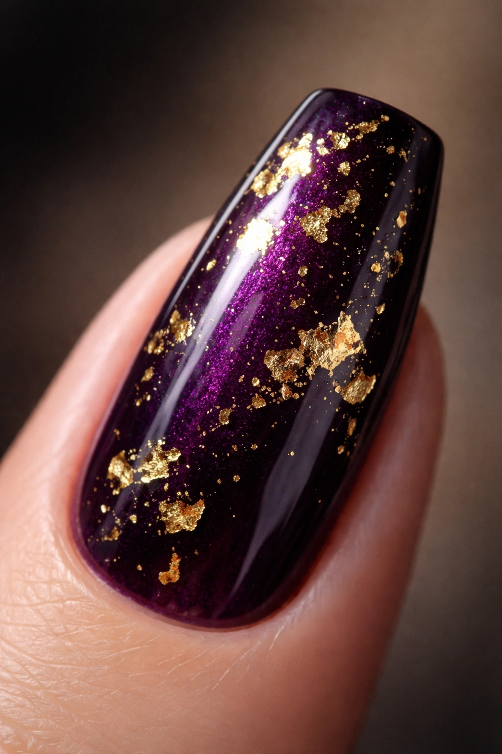 Close-up of an eggplant purple coffin nail with irregular gold leaf flecks on a neutral background