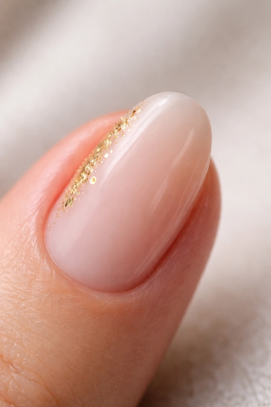 Close-up of a short almond nail with tiny gold foil near the cuticle on a nude milky base
