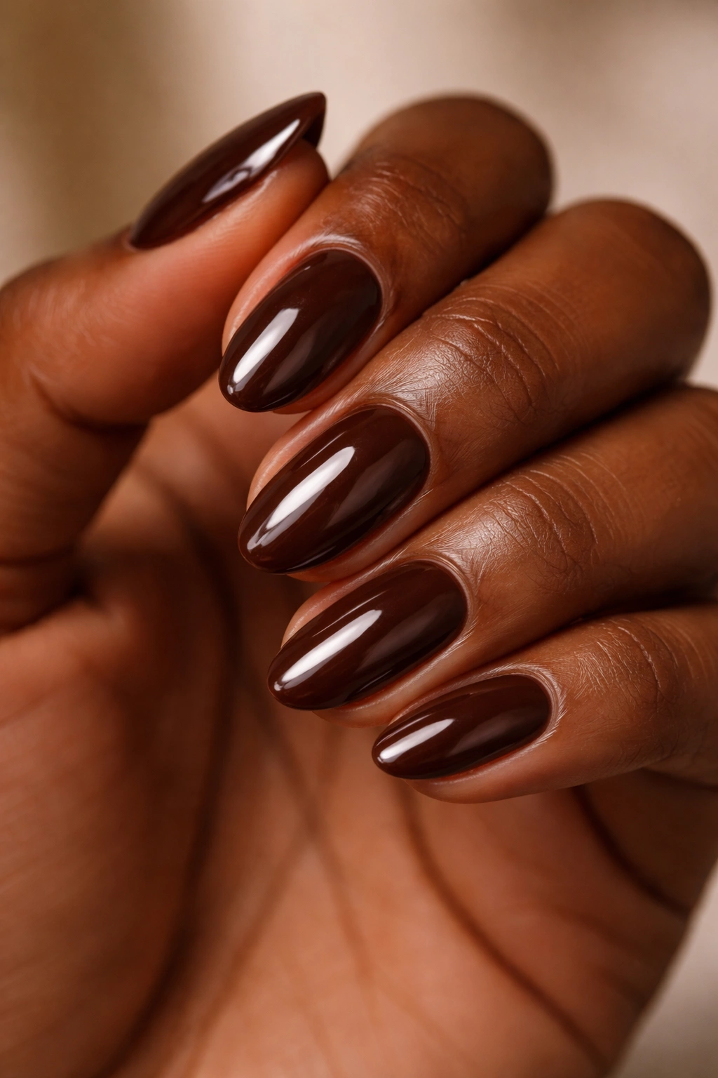 Close-up of glossy chocolate brown short almond nails on a hand with warm lighting
