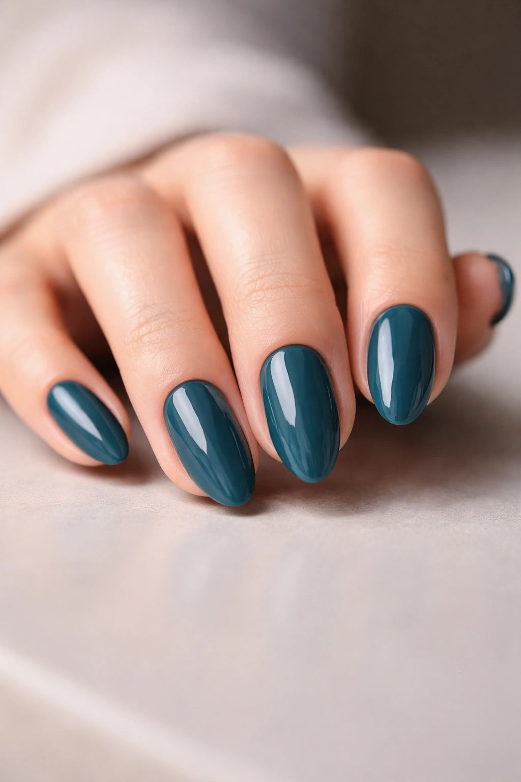 Close-up of short almond nails with glossy deep teal polish on a neutral surface