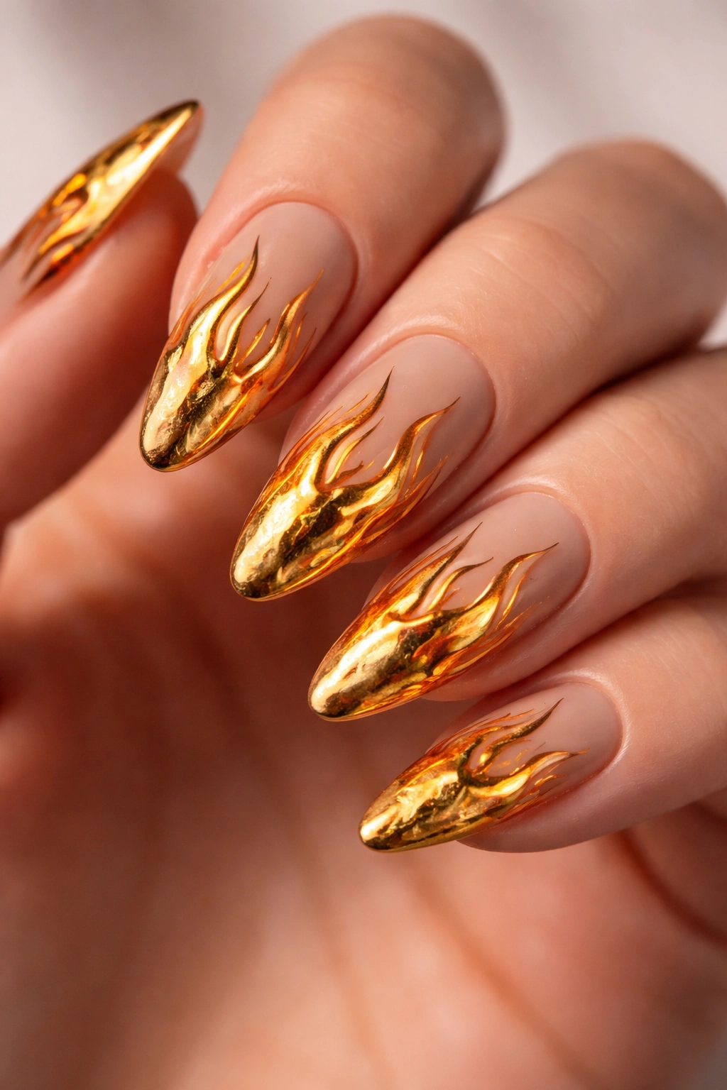 Close-up of almond nails with nude base and gold chrome flame design