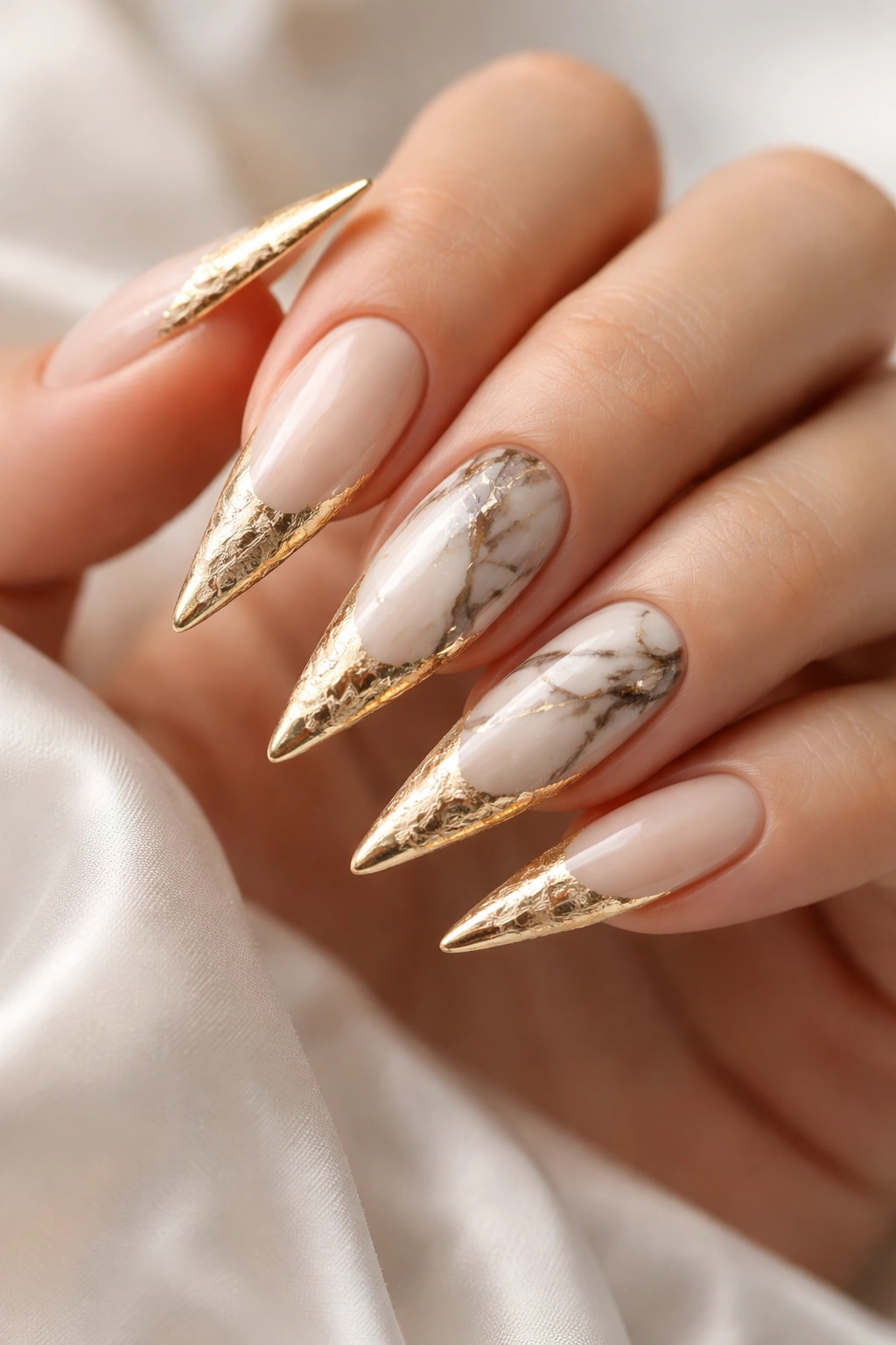 Close-up of stiletto nails with gold foil tips and marble accents on a cream base.