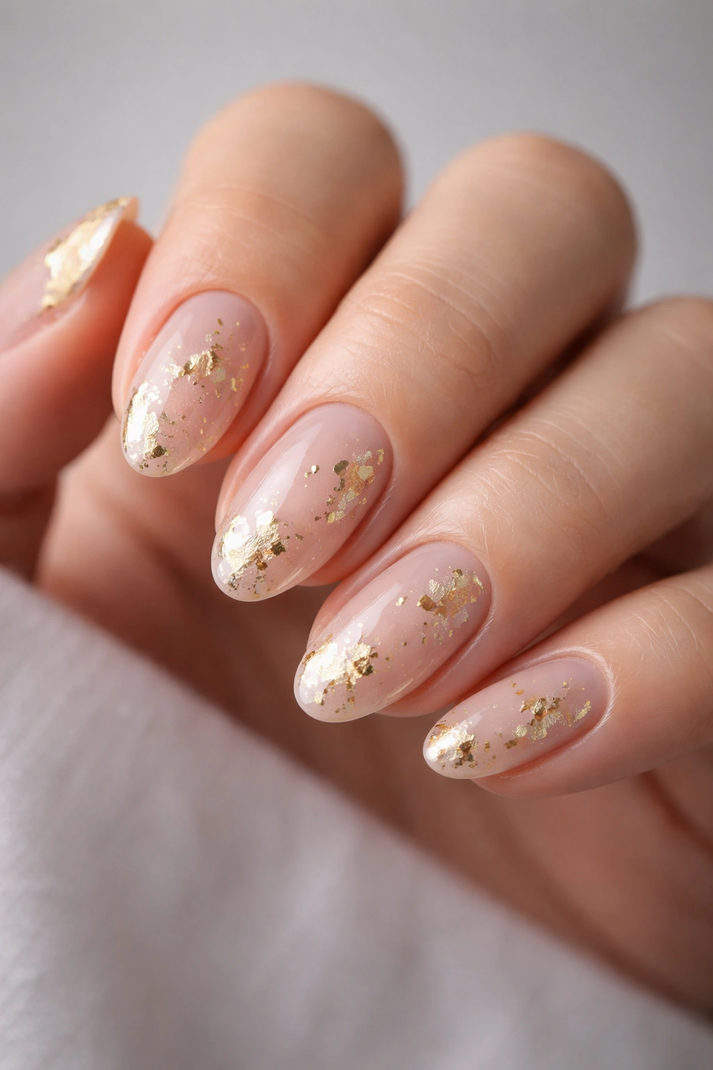 Close-up of short almond nails with sheer nude base and irregular gold foil accents.