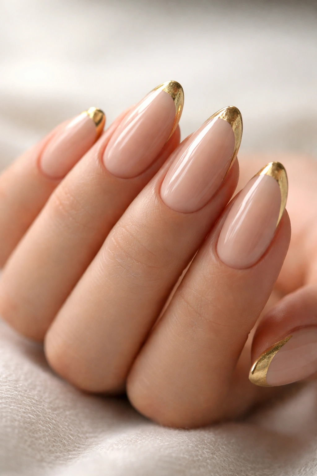 Hand with pristine almond nails featuring gold French tips on nude polish