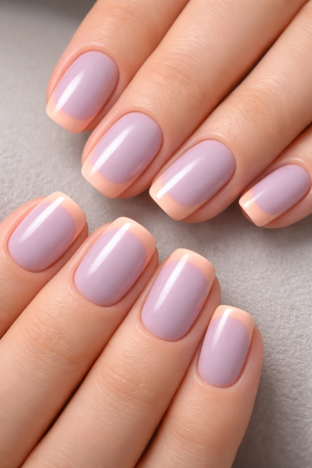 Close-up of short squoval nails with dusty lavender base and peachy tips on a neutral surface