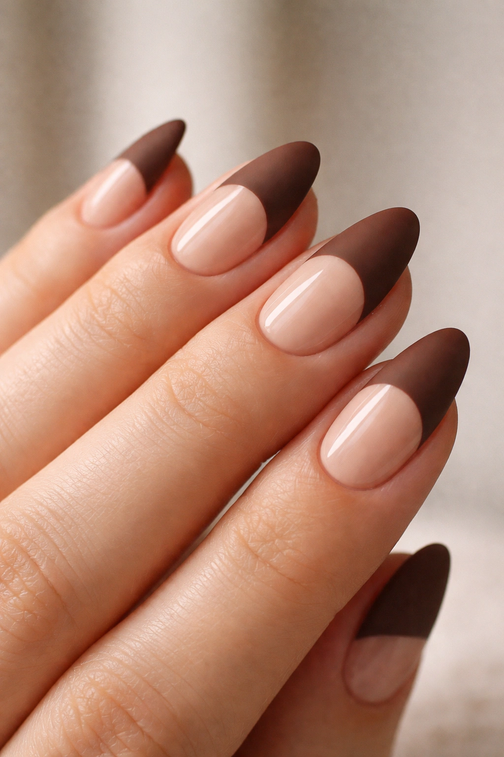 Close-up of a hand with short almond nails showing matte brown tips on a glossy nude base