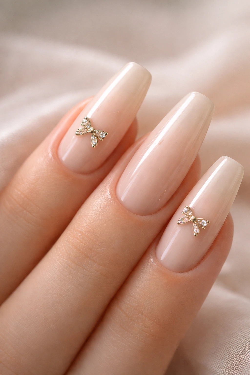 Close-up of milky beige ballerina nails with tiny metal bow charms on a neutral background