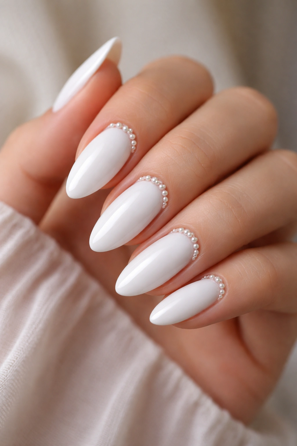 Close-up of glossy white almond nails with tiny pearl accents near the cuticle on a hand