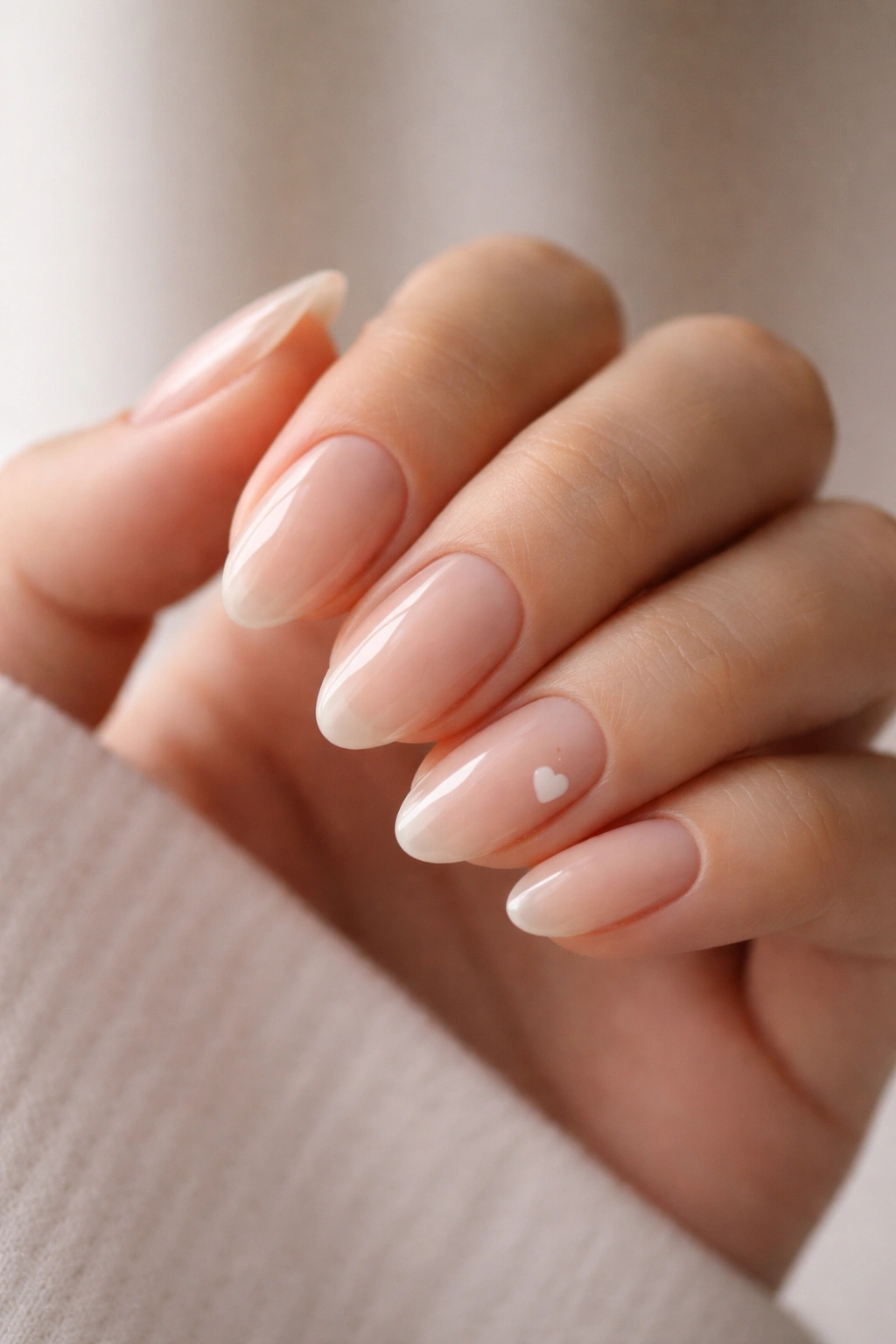 Close-up of short almond nails with nude polish and a tiny white heart on an accent nail