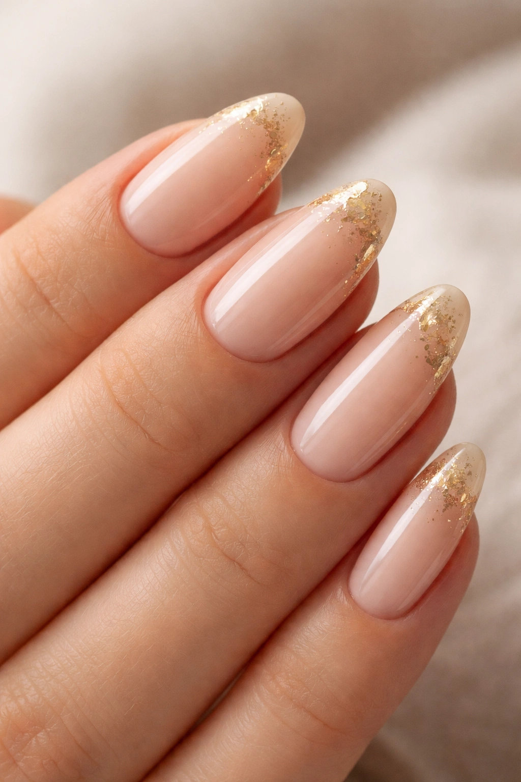 Close-up of nude almond nails with gold foil accents on a neutral background