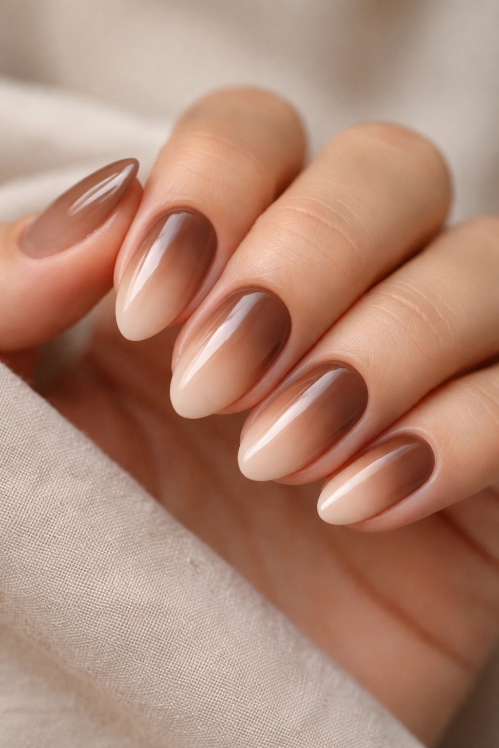 Close-up of short almond nails in cocoa brown to nude ombre on a neutral background