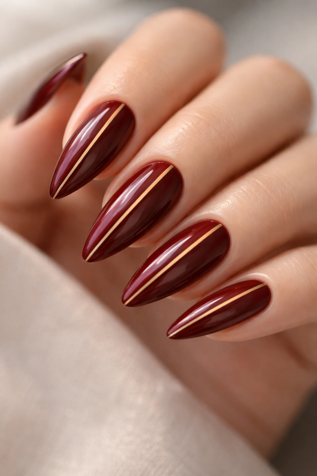 Close-up of oxblood red stiletto nails with thin gold stripes on a neutral studio background