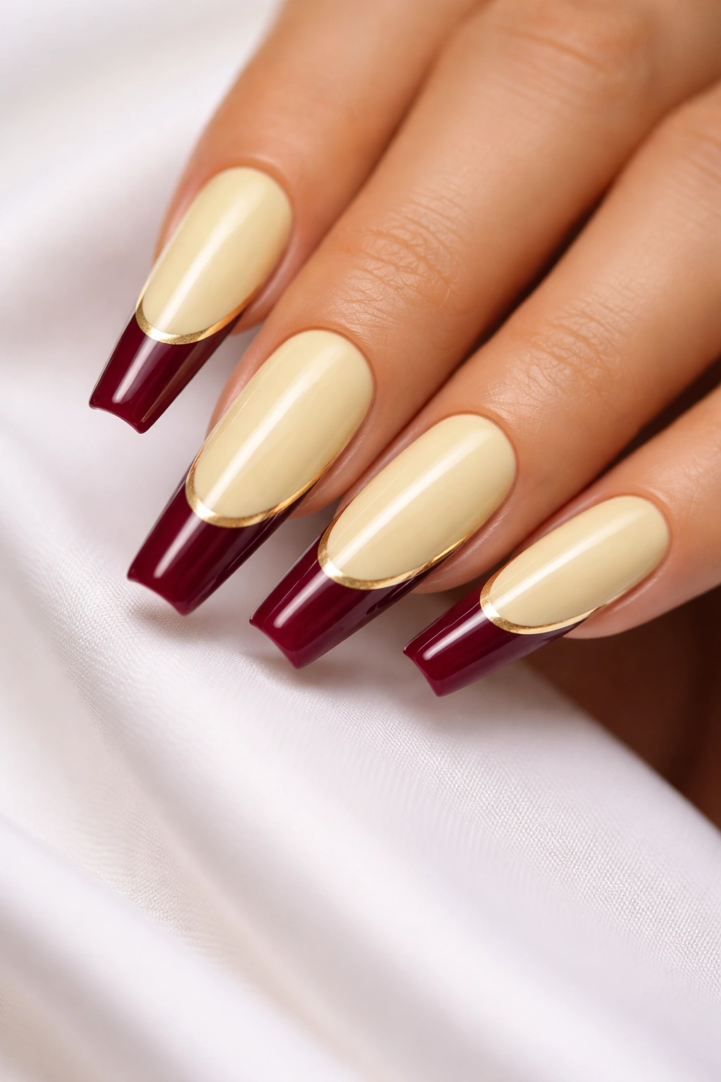 Close-up of pale yellow coffin nails with burgundy tips and gold stripes