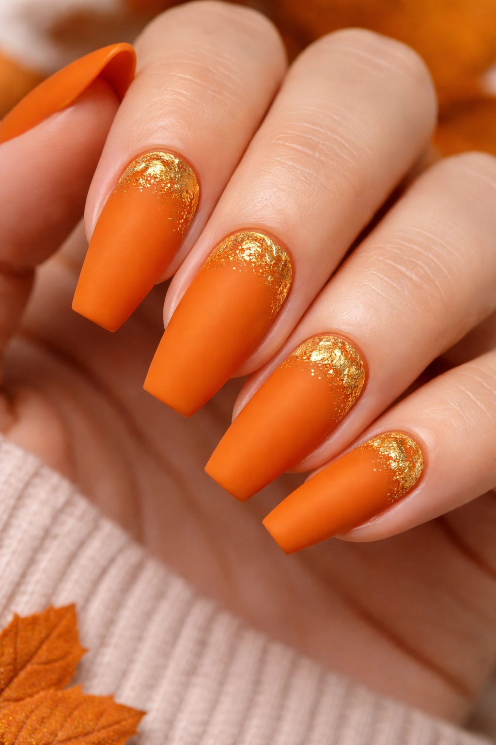 Close-up of pumpkin orange nails with matte finish and gold foil accents.