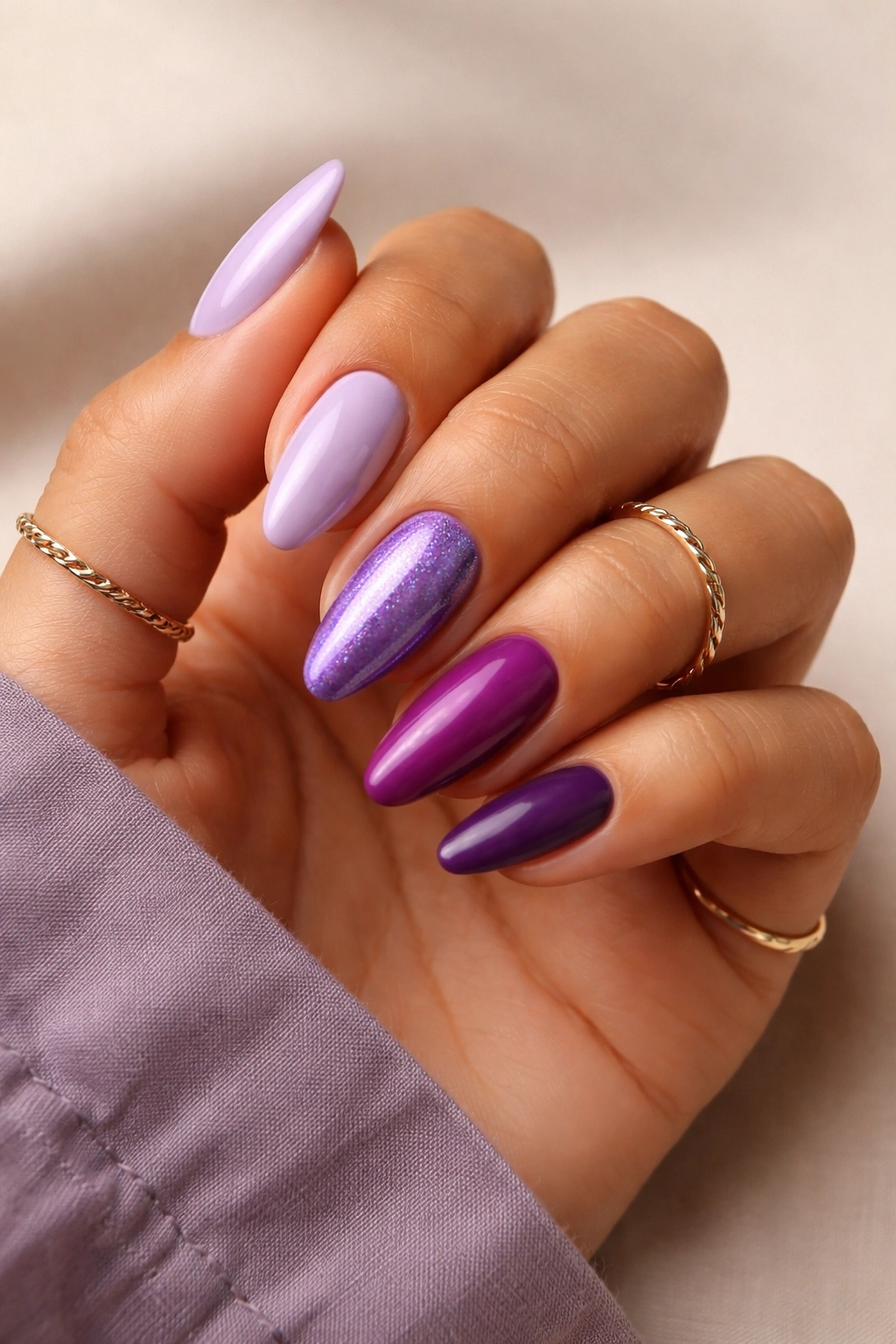 Hand with almond nails painted in multiple purple shades against a neutral background