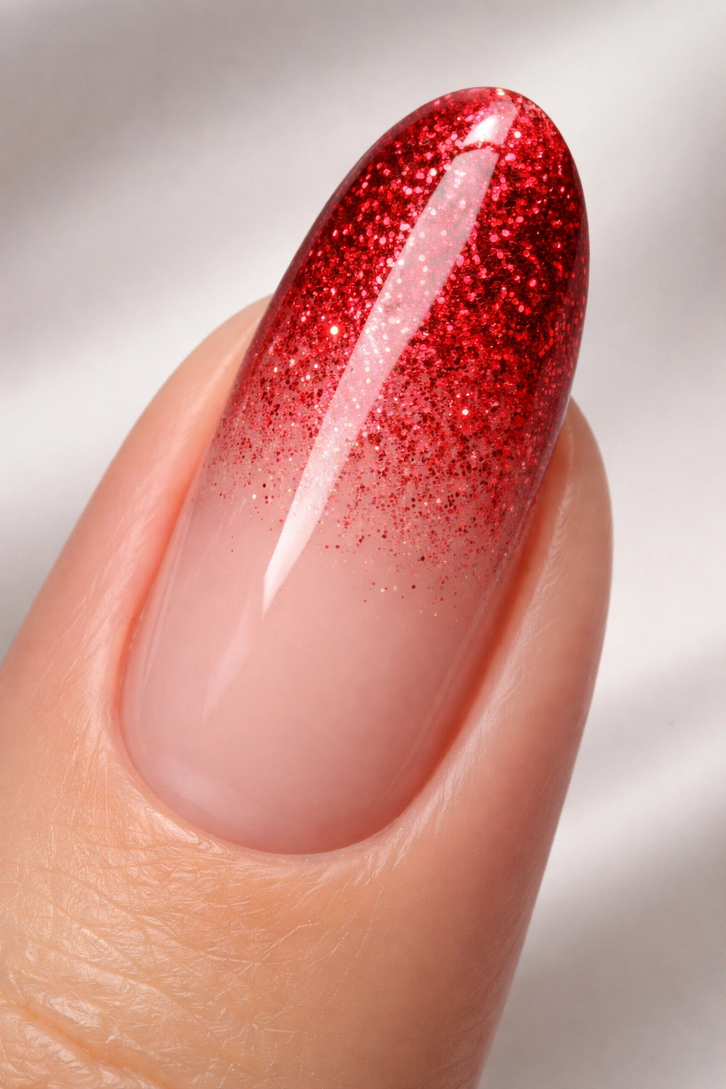 Close-up of a long round acrylic nail with a red glitter gradient fading to nude under studio lighting