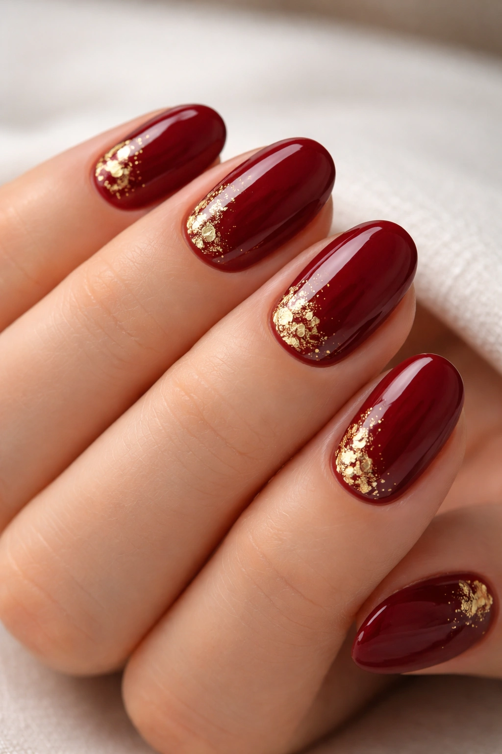 Close-up of short burgundy red nails with irregular gold foil accents near the cuticle.