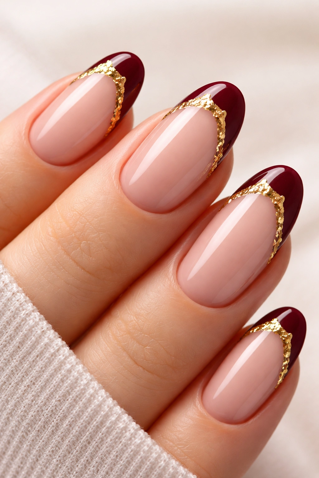 Close-up of oval nails with nude base and burgundy reverse tips accented by gold foil.