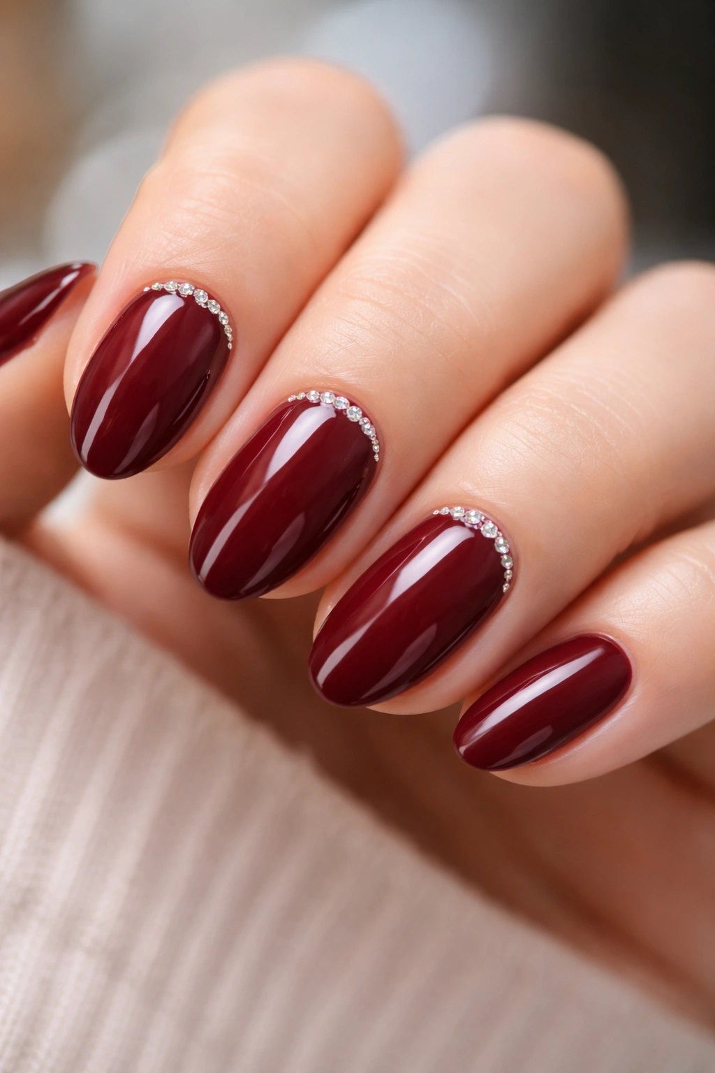Short almond nails in glossy wine red with rhinestones at the cuticle on a plain background