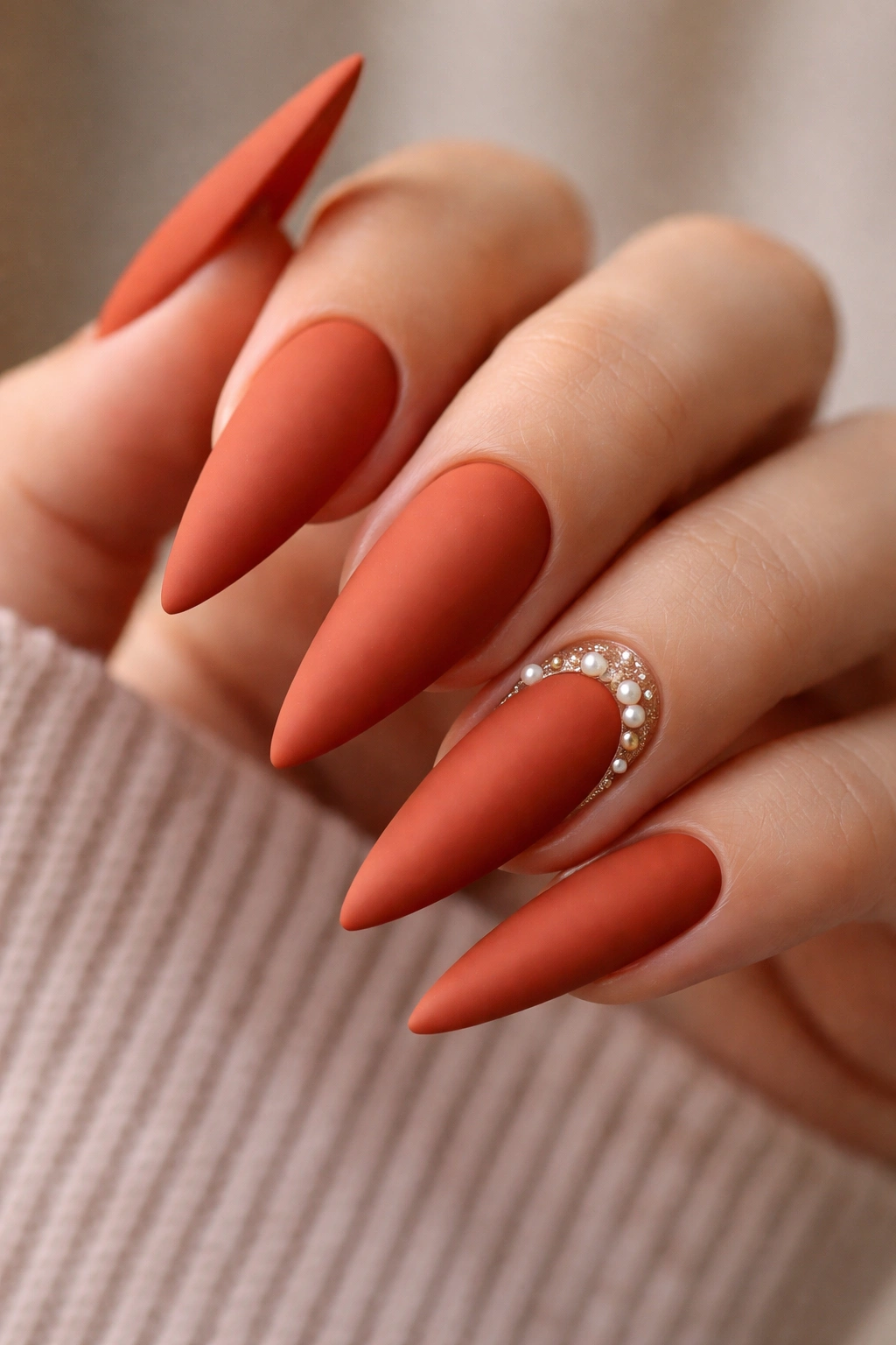 Close-up of rust-orange stiletto nails with a matte finish and pearl accents