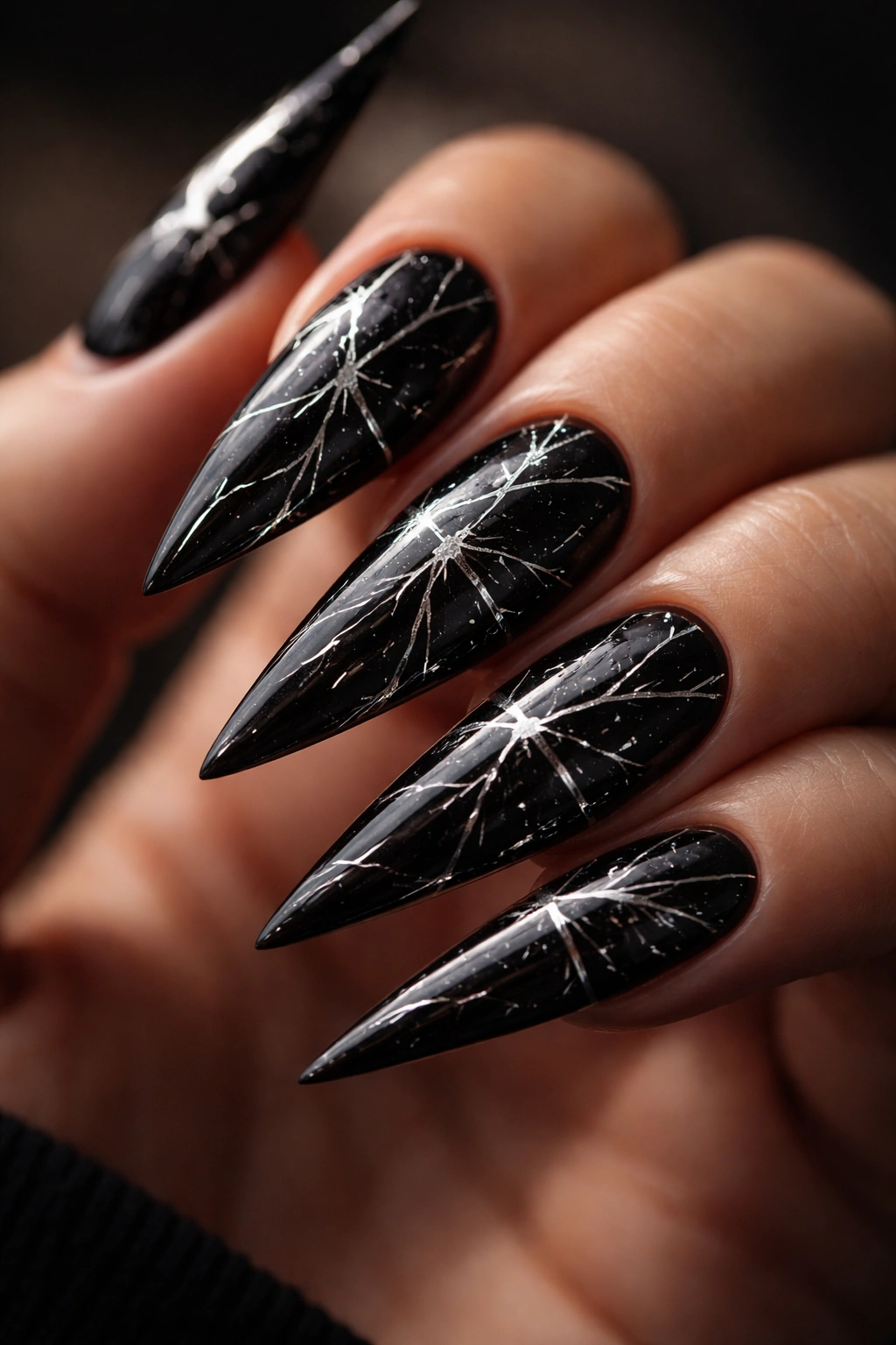 Close-up of black stiletto nails with silver and white crack lines resembling shattered glass