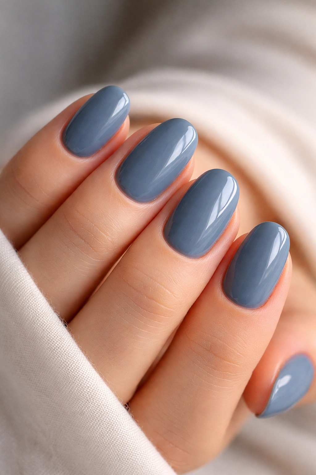Close-up of slate blue short oval nails with a glossy finish on a neutral backdrop