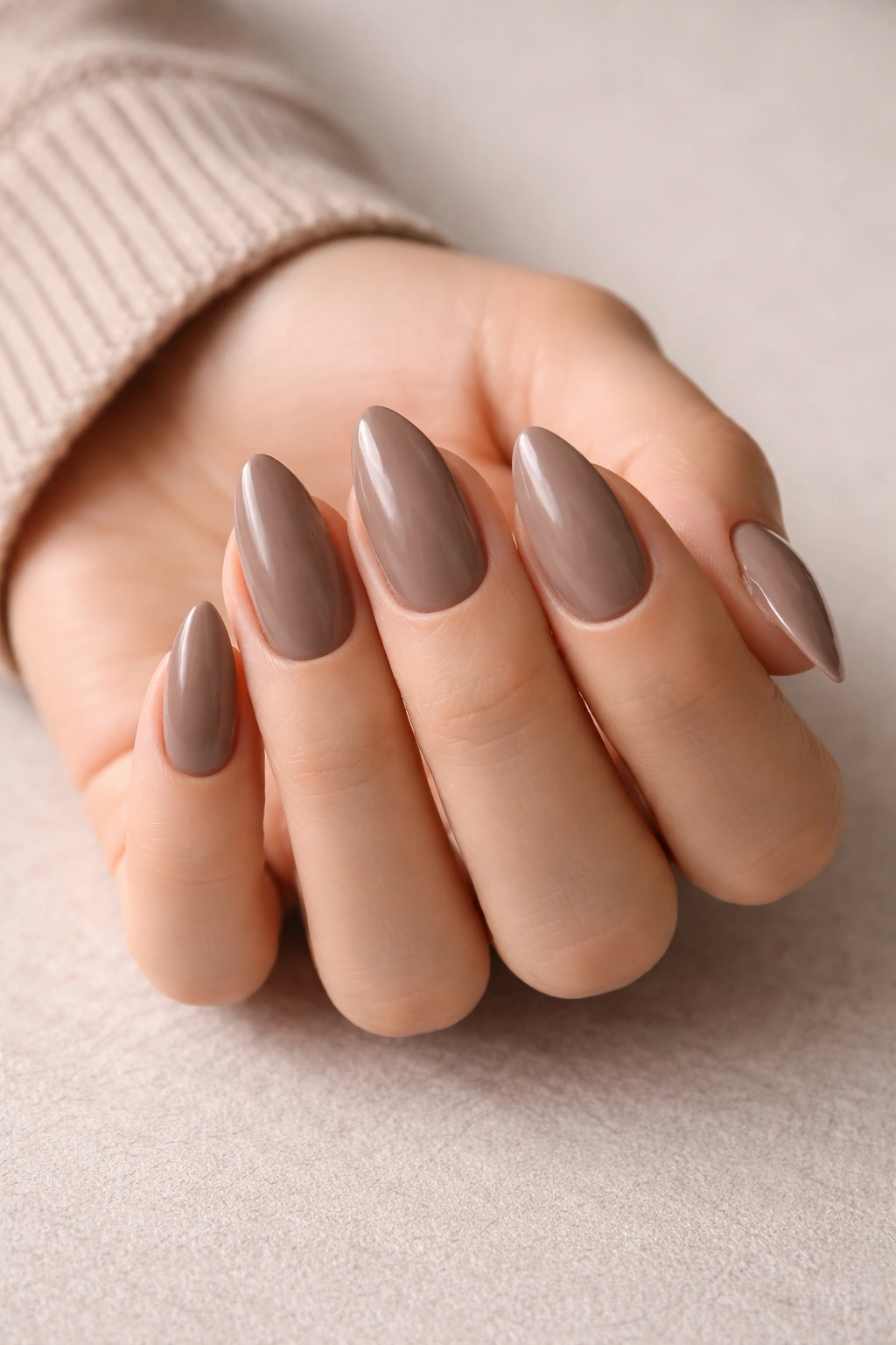 Close-up of taupe-brown almond nails with glossy finish on a neutral background.